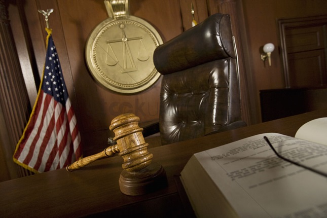 Judge's stand with a chair, gavel, book open in half in the front. Behind is an American flag and a Scales of Justice picture on the wall
