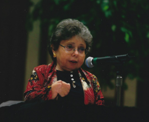 A white woman with glasses and dark grey hair, dressed in a red floral cardigan over a black blouse, speaks into a microphone at a podium.