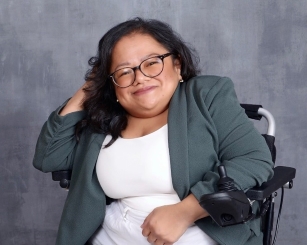 A woman using a wheelchair with glasses and black hair, dressed in gray blazer over a white blouse, in front of a gray background.
