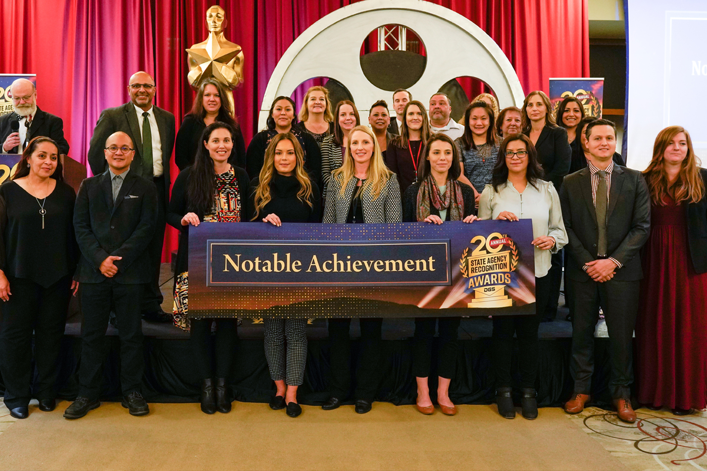 Representatives from the departments that won the Notable Achievement award pose for a group photo on stage with a "Notable Achievement" banner.